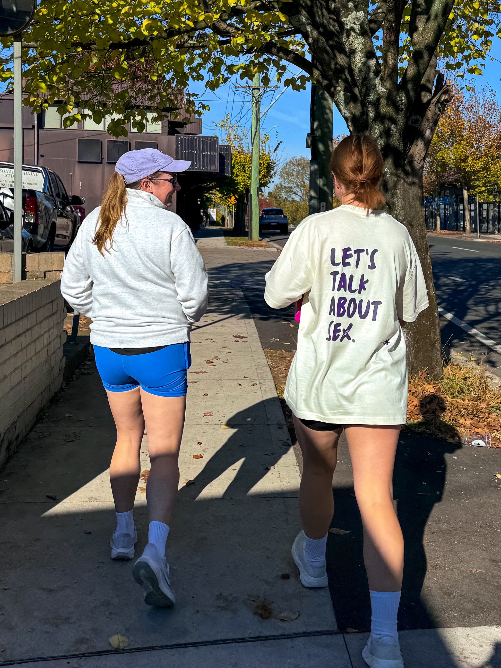 Two people walking on a sidewalk, one wearing a white shirt that says: Let's talk abou sex.